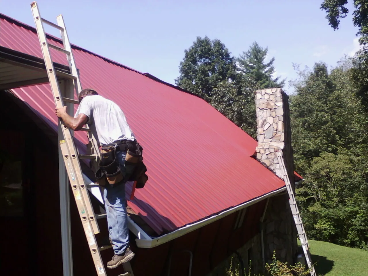 Roofer on a ladder beside a red metal roof and stone chimney — Soffit & Fascia Repair by Roofing Brothers in Owosso